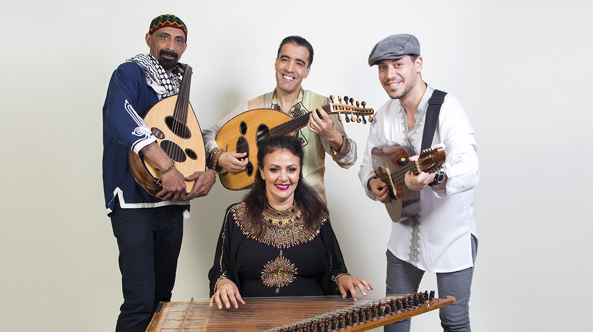 Two male musicians holding ouds stand next to a guitarist, while a female musician sits behind a qanun, all wearing North African dress.